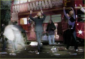 A nighttime street scene with a red double-decker bus in the background. Several people with their arms raised are standing near the bus amid scattered debris/litter on the ground, suggesting some kind of commotion or chaotic event.