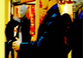 Blurry mannequin-like figures with smooth, featureless faces standing near a wall of red-lit displays. A dark-clothed figure is prominent in the foreground.