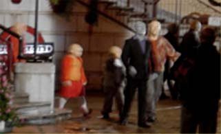 A blurry street scene with several human-like plastic figures (mannequins) standing near what looks like a shop window or arcade display. There's a group of figures clustered together — a child-sized figure in an orange top, a couple of taller adult figures in dark suits and casual jackets, and others in the background — all with the smooth, featureless look typical of shop-window mannequins. The setting appears to be an urban street or shopping area with stone paving and a building facade with windows in the background.