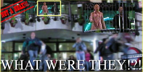 Collage-style image featuring a blurry nighttime photograph outside a mall, with several people running in the foreground. Two inset boxes at the top highlight a pair of figures standing on an upper balcony, with one wearing a pink outfit and the other dressed in dark clothing. A large red diagonal stamp in the upper-left corner reads 'NOT A HOAX!!!' and oversized white text across the bottom asks, 'WHAT WERE THEY!?!'