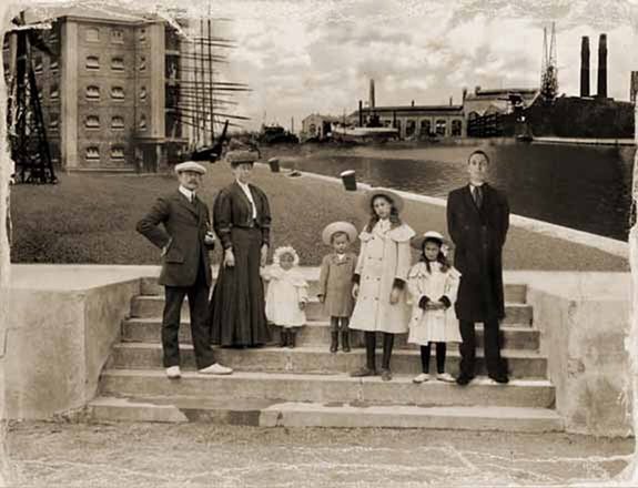 Black-and-white family photograph taken in Southampton in 1912, showing seven people standing on stone steps beside a dock. An adult couple stands on the left with four young children between them and a tall man on the far right, identified as the Doctor. Behind the group are a waterfront, sailing ship masts, warehouses, industrial buildings, and factory chimneys, reflecting the busy port environment of early 20th-century Southampton.